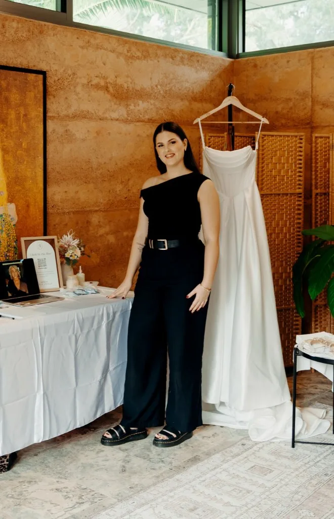 Holly Amelia standing beside a hanging bridal gown, wearing a black one-shoulder top and wide-leg trousers