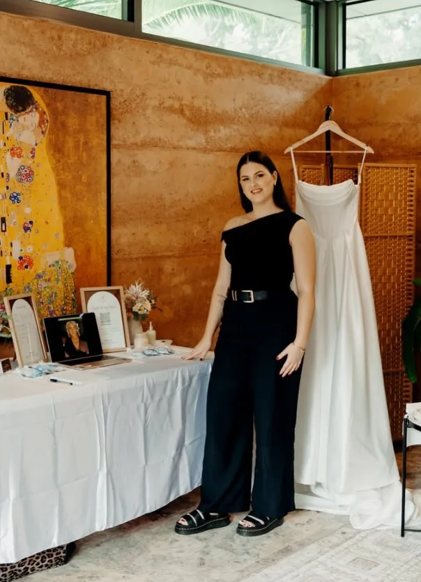 Holly Amelia standing beside a hanging bridal gown, wearing a black one-shoulder top and wide-leg trousers